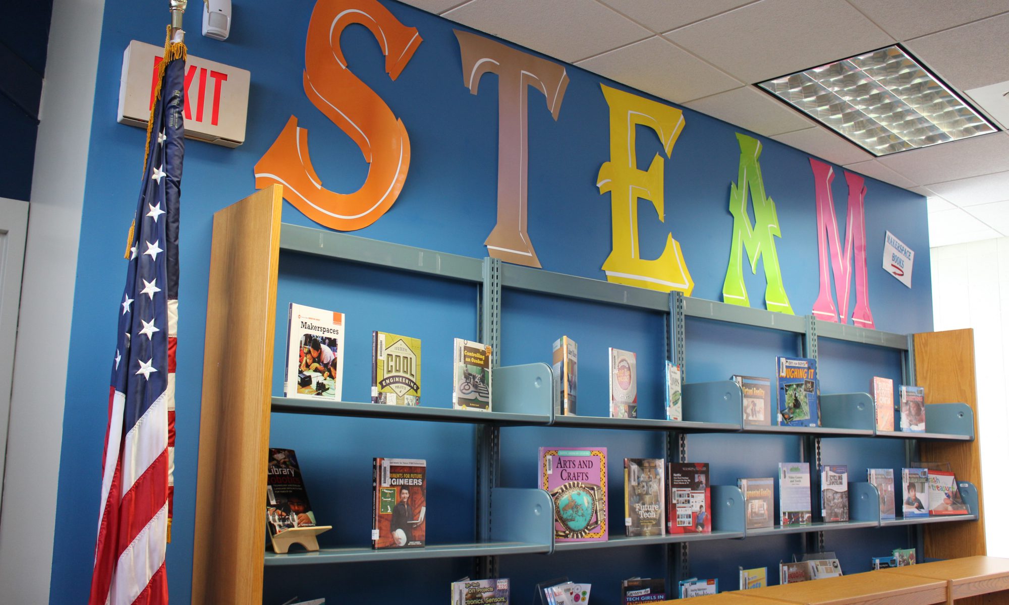 Book shelves withe an American flag to the left of it and very large letters spelling STEAM above them.