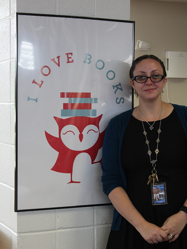 Woman in black dress and glasses, long silver necklace stands in front of a poster that says I Love Books