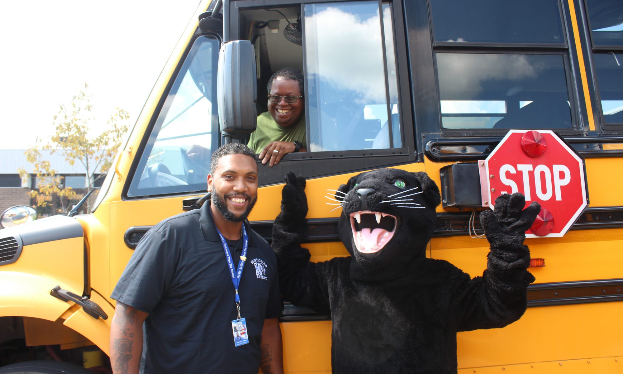 A bus driver in a school bus smiling with panther mascot and another Monticello employee.