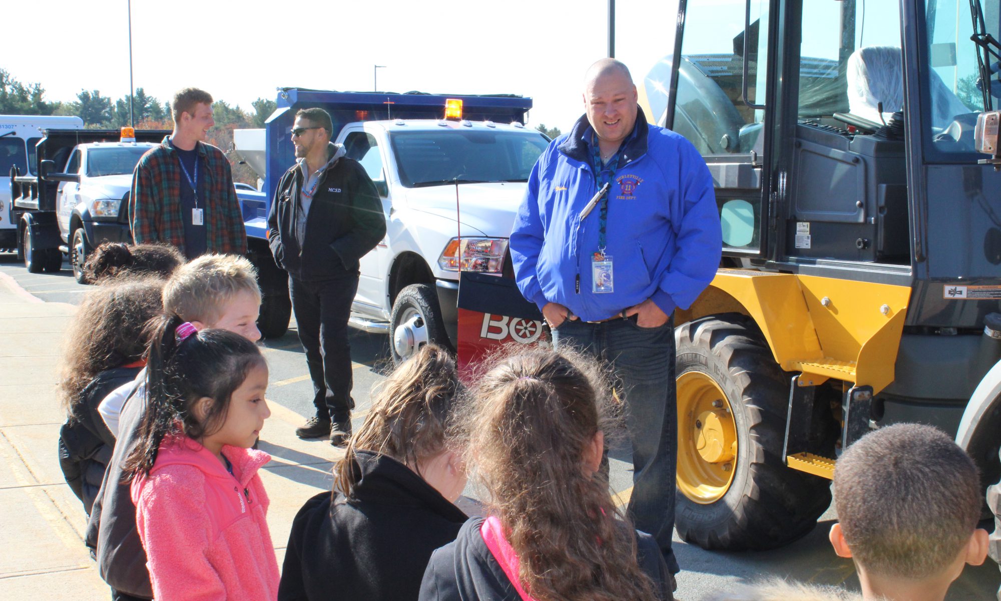 A man in a blue winter jacket speaks to a group of elementary students about facilities. There are many different trucks behind him.