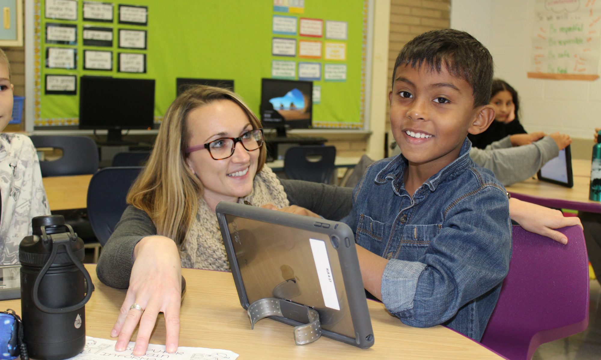 A young boy smiling, wearing a blue shirt, as a teacher helps him with his electronic device.