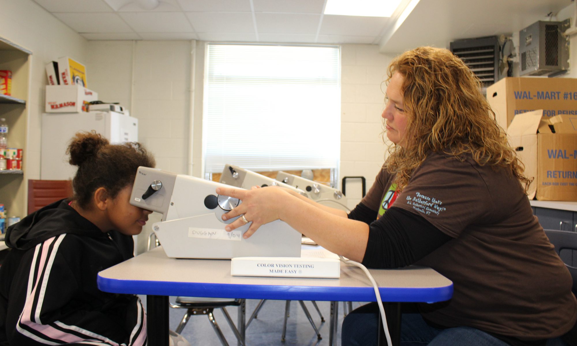 An elementary student looks into a device sitting o the table that will measure her eye health. A technician with a brown t shirt on and shoulder-length blond hair administers the test.