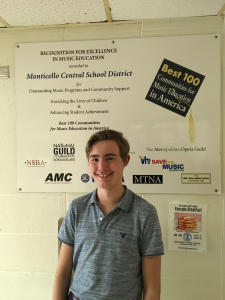 High school student, Edward Spear, wearing a blue short sleeved shirt stands in front of the Best 100 Communities for Music Education in America banner.