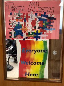 Posters on the door of a classroom showing a puzzle made up on different flags and another poster with a rainbow of colors for a background saying Everyone Welcome Here.