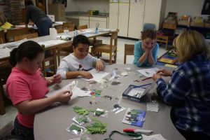 three students sit around a table while a teacher helps them with school work 