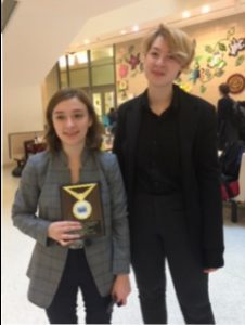 Sam Leoniuk and Bailey Yewchuck stand next to each other and smile holding the plaque and trophy that they received at the debate tournament 