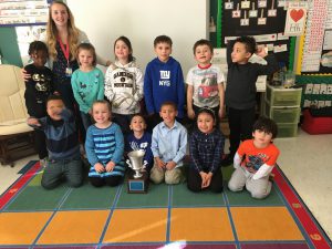 Mrs. Moore and her kindergarten students sit together on a rug smiling