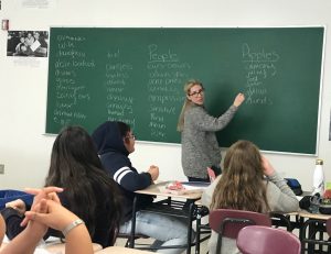 Teacher writes words on the blackboard while girls watch. The words describe people and fruit.