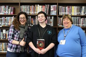 Two women flank a high school student who holds a book. Woman on left is giving a thumbs up.