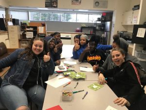 a group of students sit around a table and smile as they write cards.