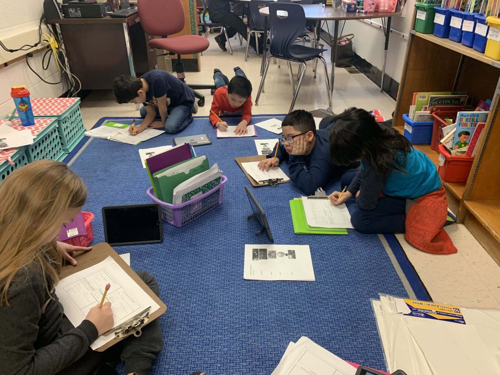 Five elementary students sit on a blue rug, each with a chromebook, clipboard and pens writing stories.