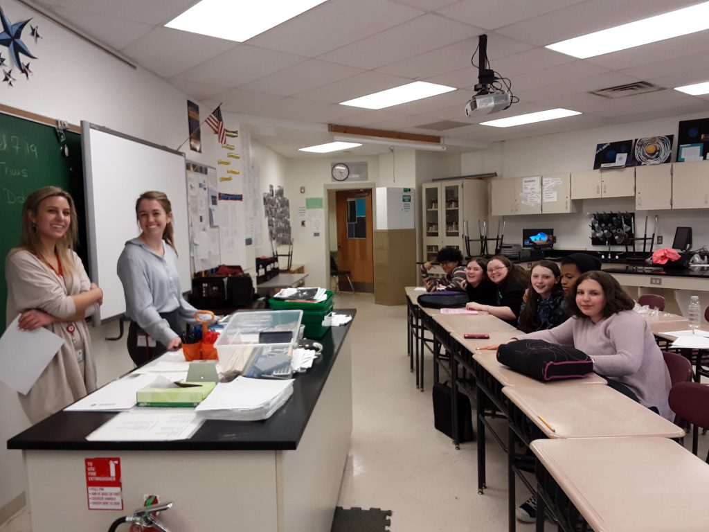 In a classroom setting, two women speak to a group of  six middle school students.