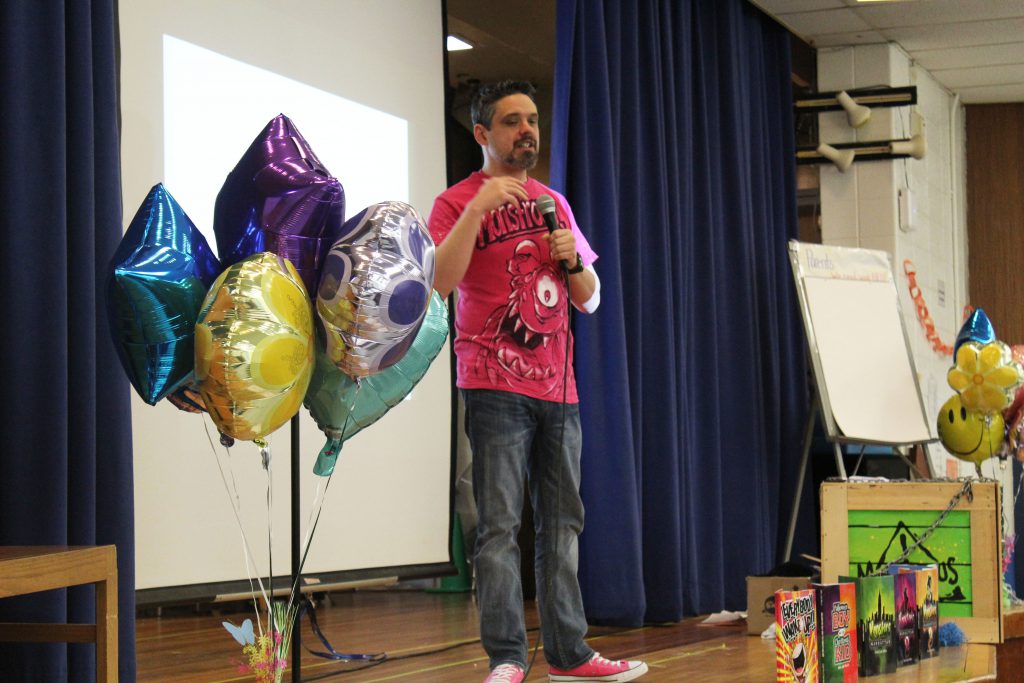 The author stands on a stage with balloons to his left speaking to the students