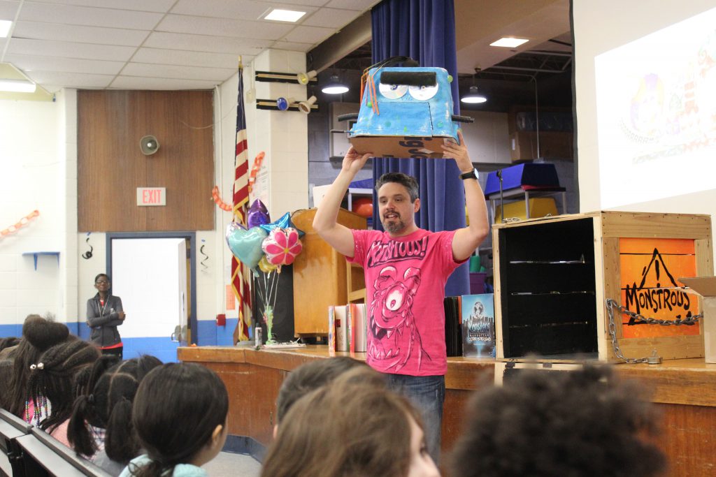 The author, a man in a pink shirt, holds up a blue monster he created