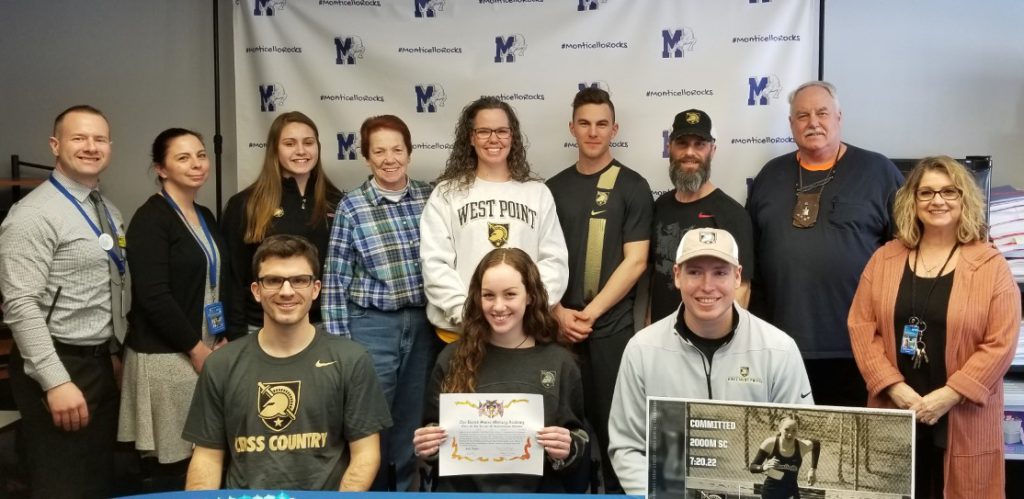 High school girl holding up a document she just signed. She is flanked by her coaches and behind her are nine people, her family and administrators