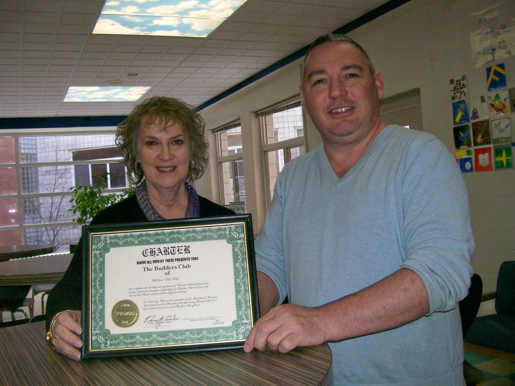 A woman and man are smiling. he woman presents a club charter from 1990 to the man, who is the club's advisor.