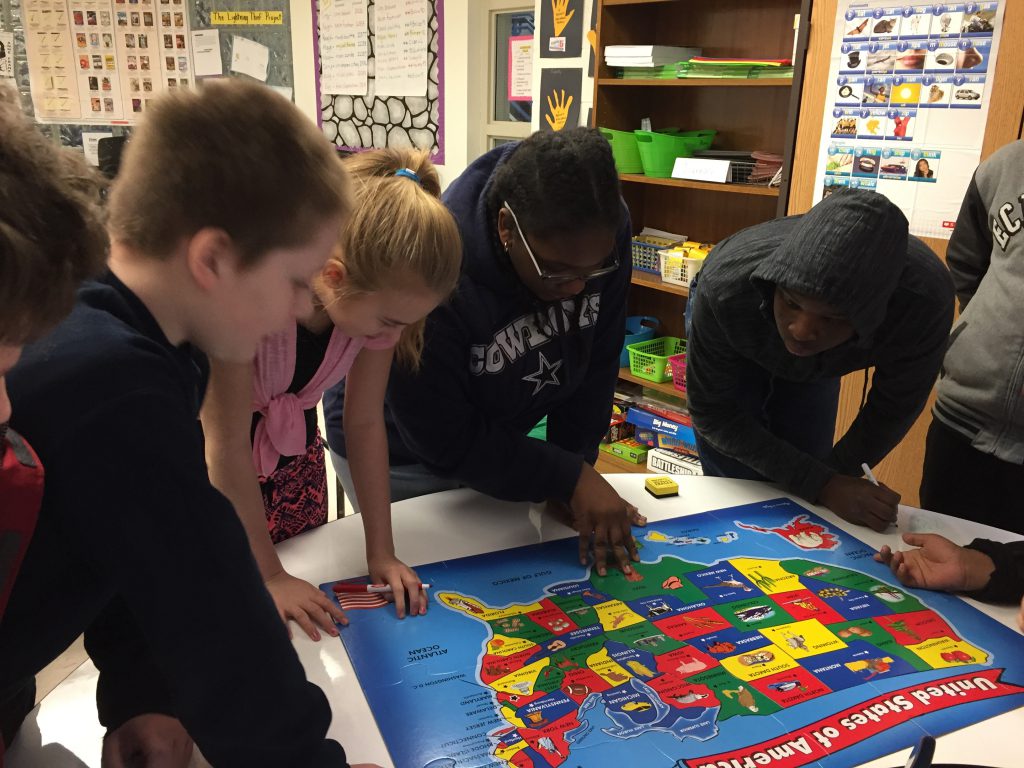 Four middle school students are bent over a table completing a puzzle of the United States.