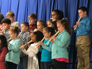 Group of elementary students on stage with blue curtain behind them play their recorders