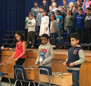 On stage elementary students sing while three play xylophones