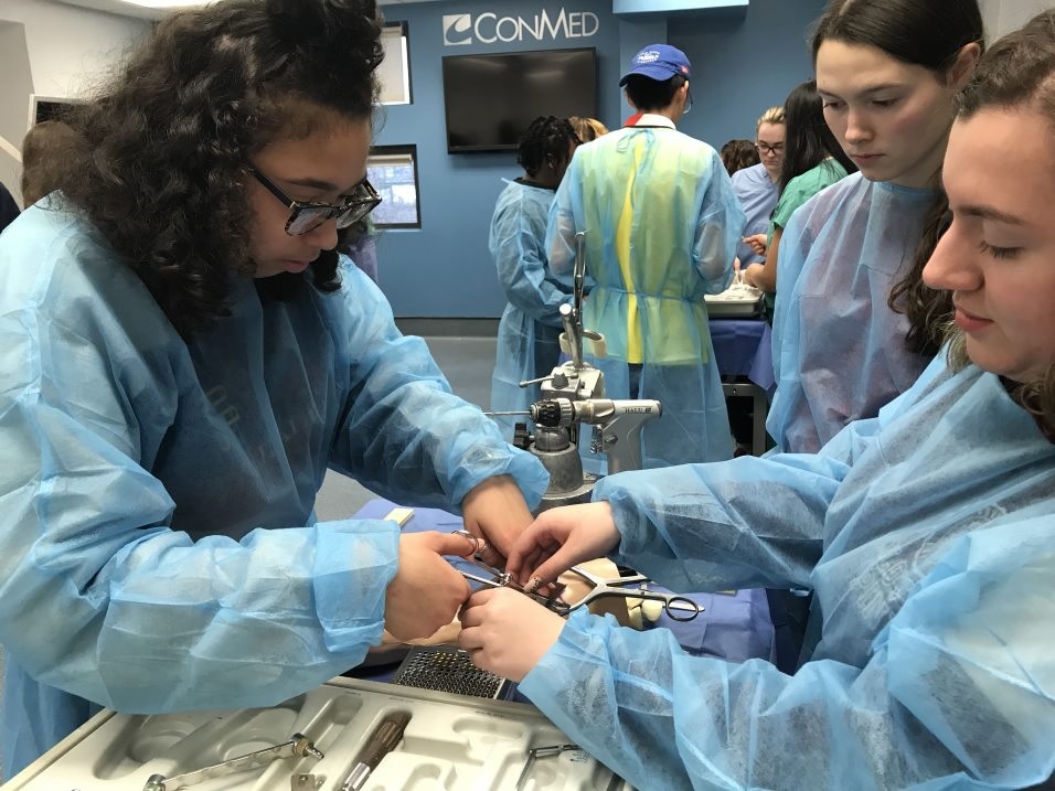 Three students dressed in blue operating gowns are operating on the simulated hand. 