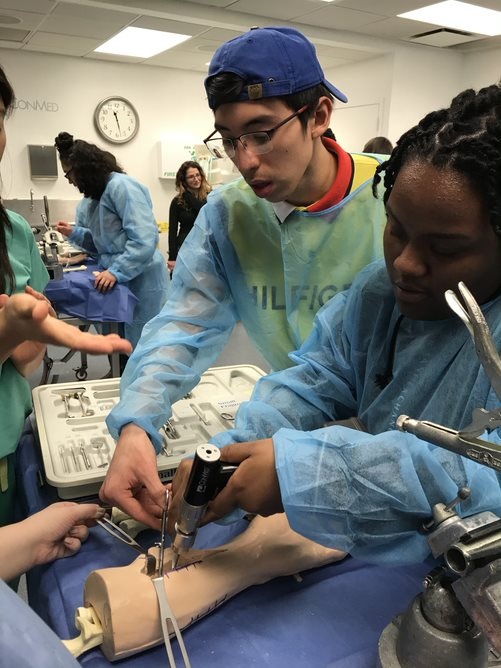 Two students work on the simulated arm, one holding back the skin with an instrument and the other fixing the bone.