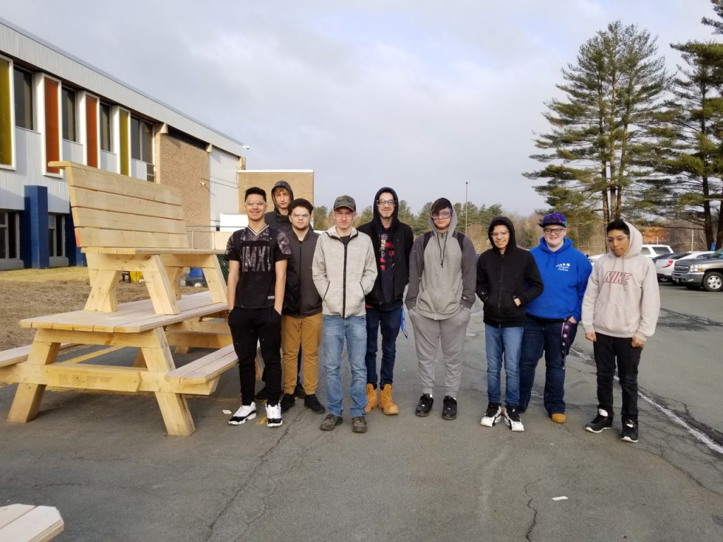 Nine high school boys stand next to a wooden bench and a picnic table they made in class.