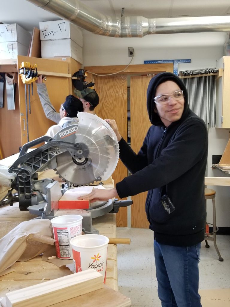 A student in a blue hoodie cuts a piece of wood using the circular saw