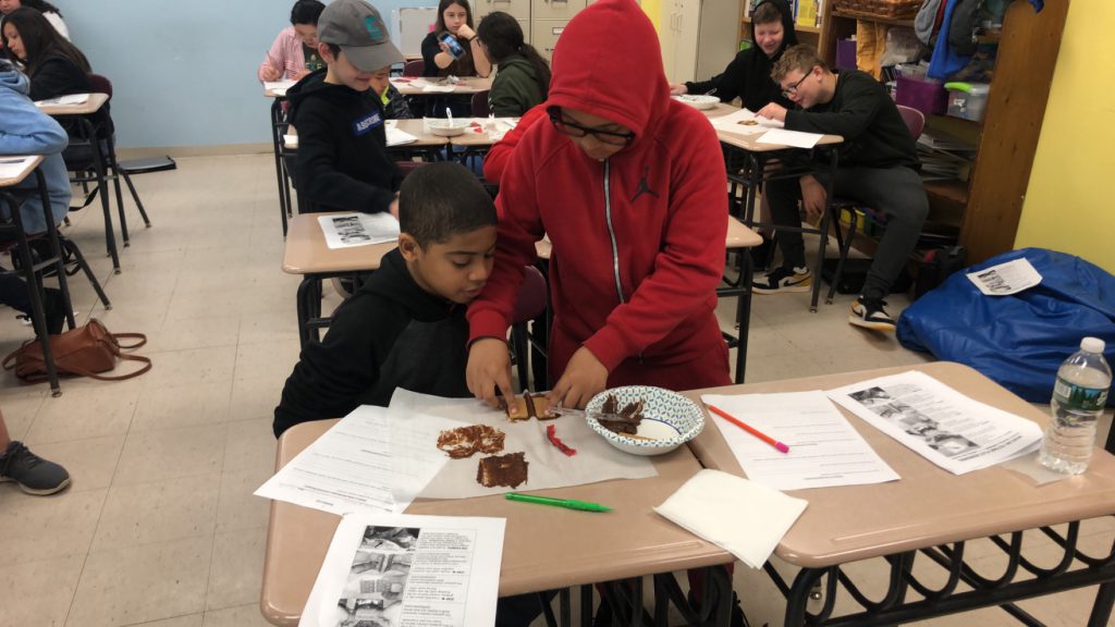 Two boys  - one in a red hoodie standingand placing items on the paper, and one sitting watching.