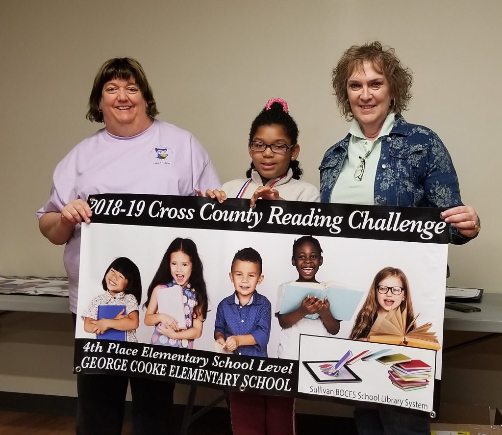 Two women and one elementary student hold a banner declaring George L. Cooke Elementary School the fourth place finisher in the Cross Country Reading Challenge