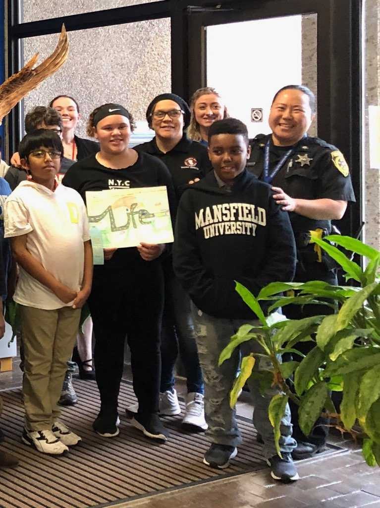 Six students and two adults, one a police officer, pose with a girl holding her poster