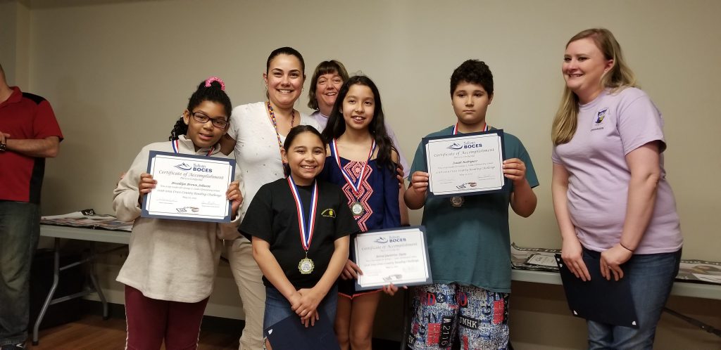 Four elementary students wearing medals hold up their reading certificates. Two adults stand behind them.