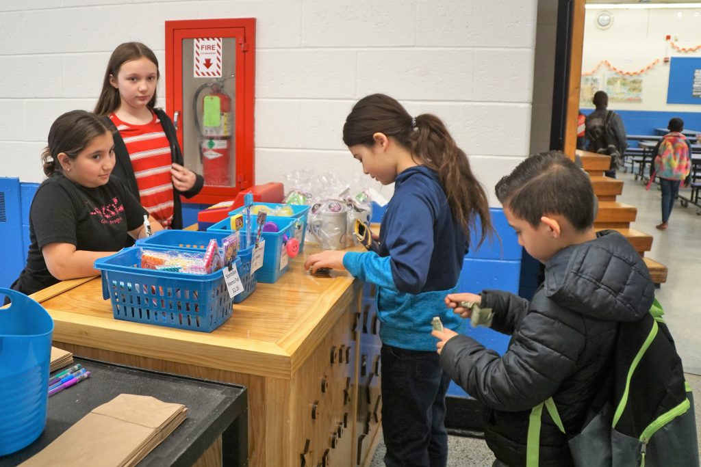 Two girls behind the store counter with supplies for sale in blue baskets on the counter. Two boys are getting their money ready to purchase supplies.