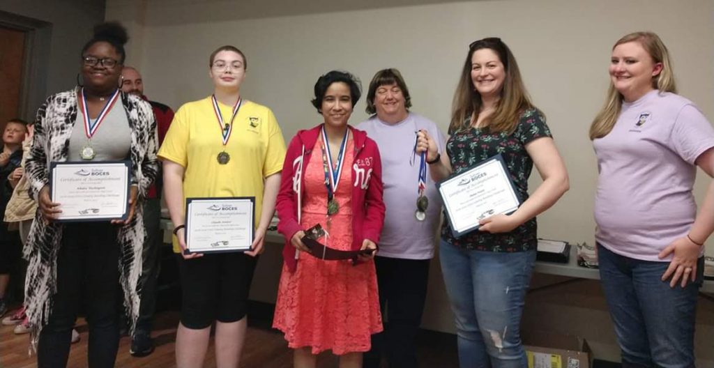 Three students wearing medals and holding certificates smile along with three adults