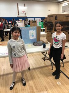 Two girls stand on either side of their science project