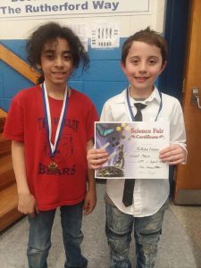 Two young students stand wearing the medals they earned. One is holding a certificate.