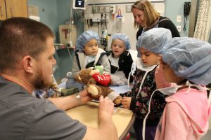 a group of students look on as a nurse helps bandage a stuffed bear 