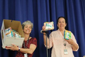 Two women smiling. one holding a box filled with pictures the other holding one of those pictures up.