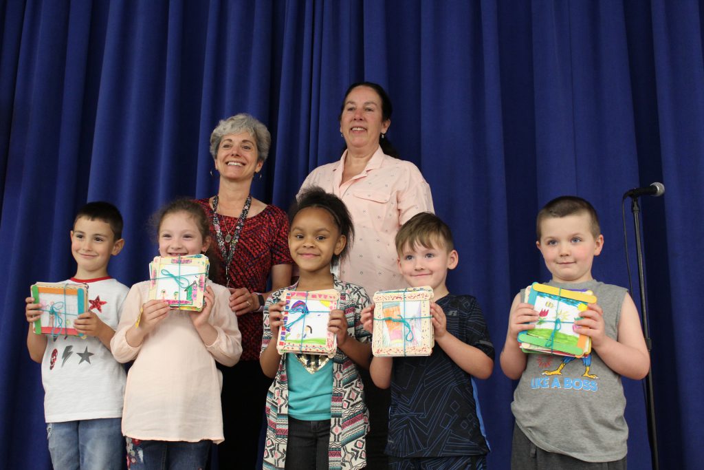 Five kindergarten students hold up pictures in front of them. Behind them are two women smiling