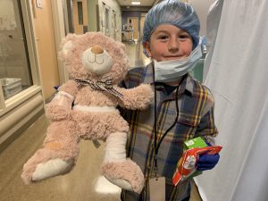 a student wearing a surgical cap and gown holds a stuffed bear in the hallway