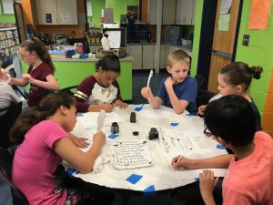 four students are sitting around a table using a quill and ink to practice writing 