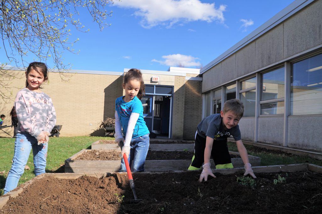 A young girl in a bright blue shirt uses a garden hoe, a boy with garden gloves on hsas his hands in the garden box dirt while another girl stands next to them with her gloves on smiling.