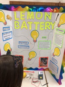 Poster board showing the words Lemon Battery in bright yellow with drawings of light bulbs all around and the experiment written out. A girl is reading it. 