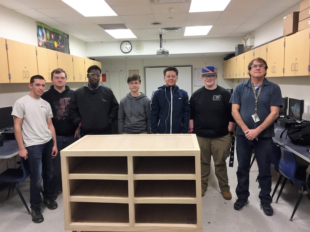 Five male students, all wearing goggles, and their teacher stand around the wooden store counter they built