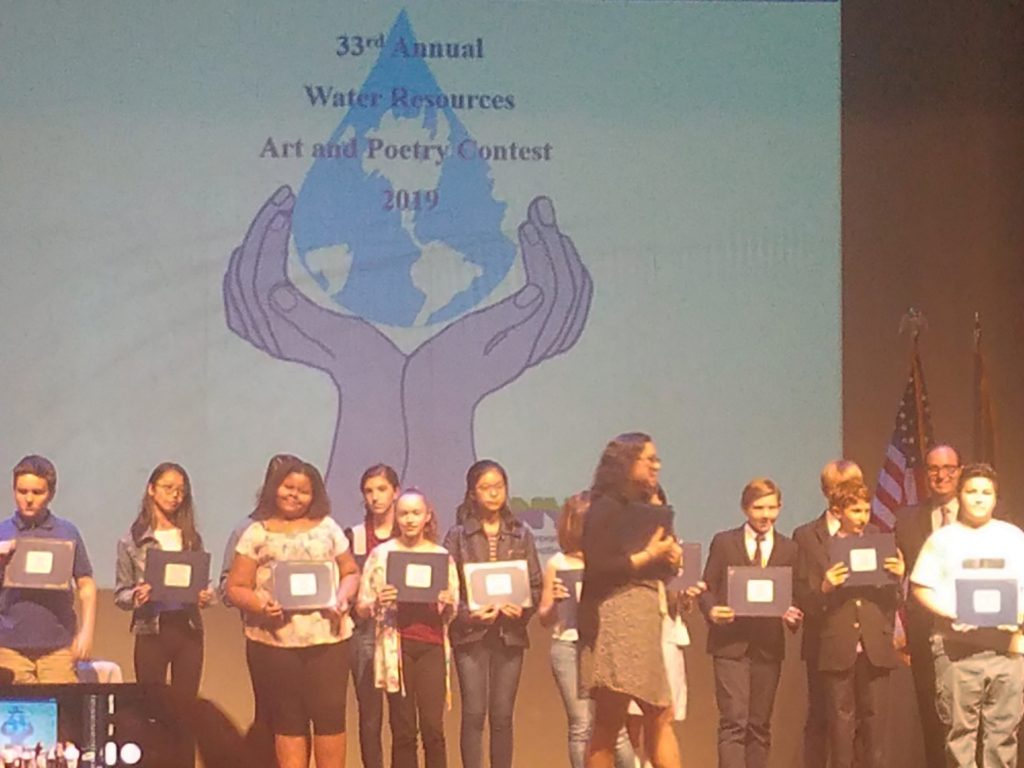 A group of students stand holding awards in front of a backdrop showing water resources