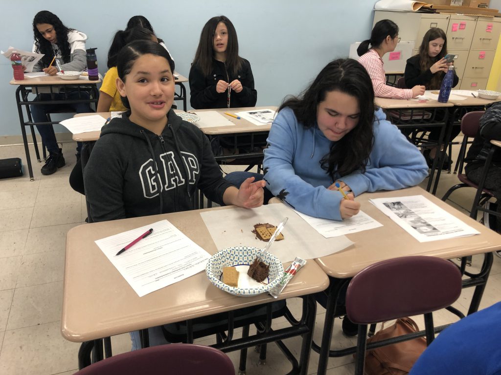 Two middle school girls sitting at desks writing and putting together their crackers, icing for the lab.