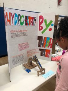 A colorful poster shows the hydro electricity experiment while a girl in a pink shirt reads it.