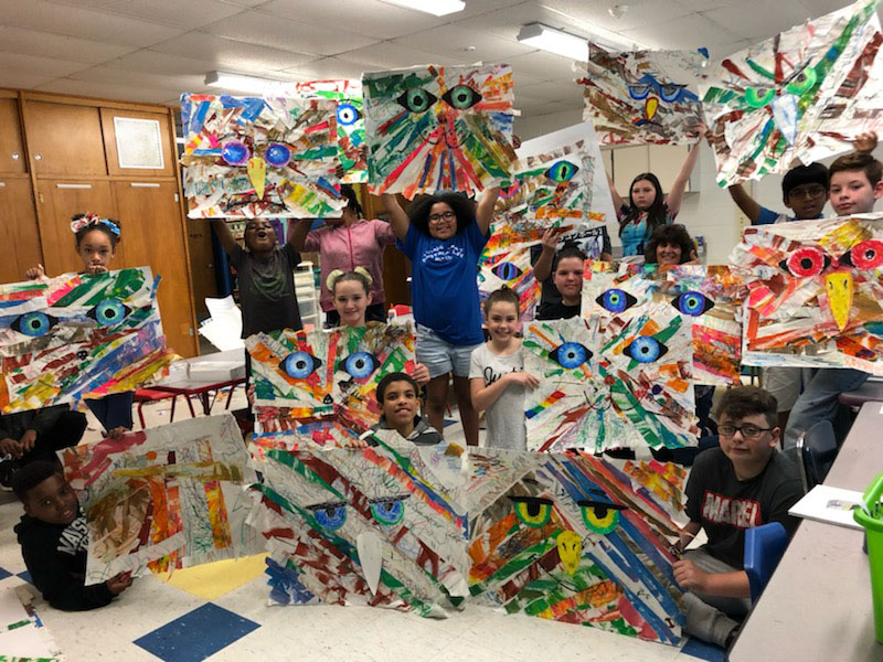 About a dozen students hold up large, multi-colored art projects