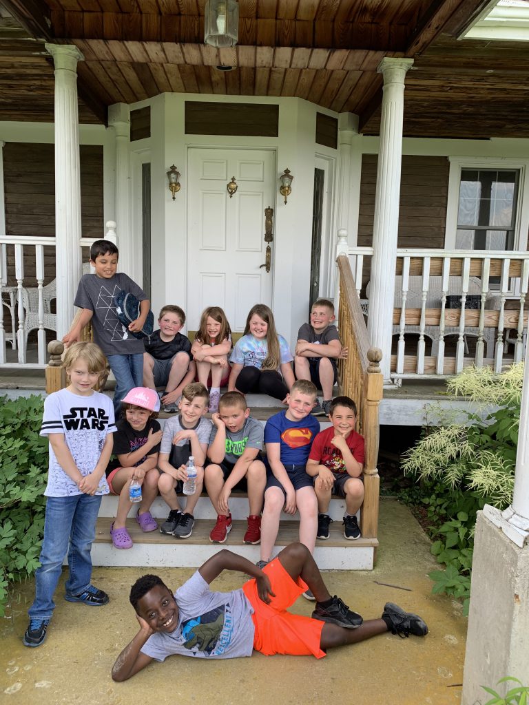 Twelve second grade students sit on the steps of a porch with one student lying across the front