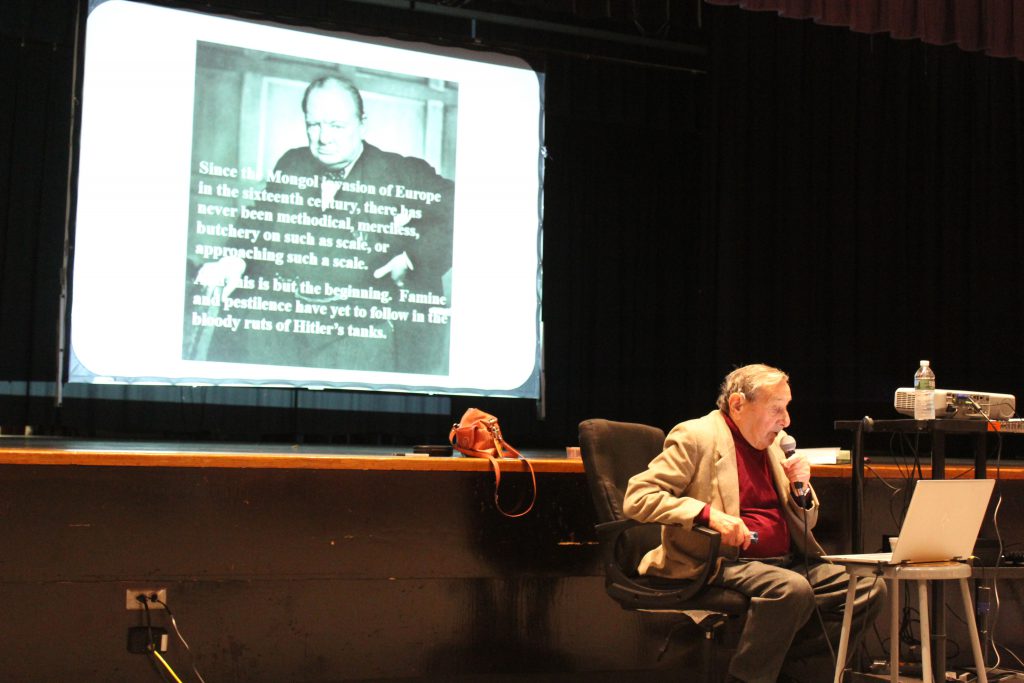 A man in a beige jacket and red shirt sits at a computer. Behind him is a slide on a large screen showing a picture of Winston Churchill.