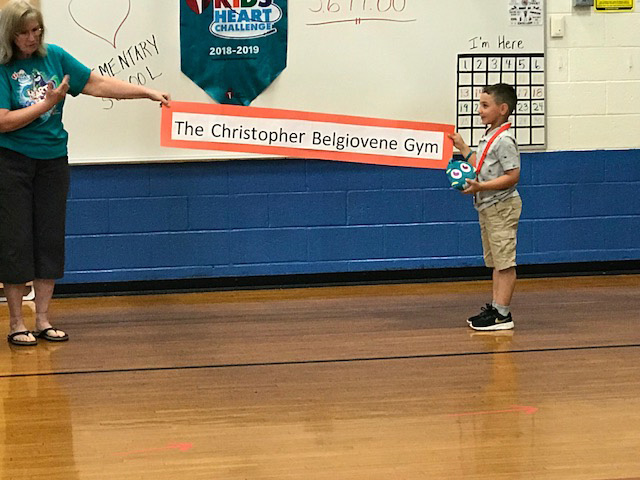 An adult holding one end of a long narrow sign and a young boy holding the other. The sign says The Christopher Belgiovene Gym.